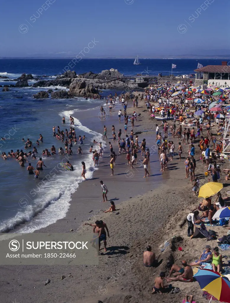 Chile, Valparaiso Region, Los Lilenes, Crowded Beach Near Vina Del Mar.  People Sunbathing And Playing In The Surf.  Rocks And Distant Yacht Beyond.