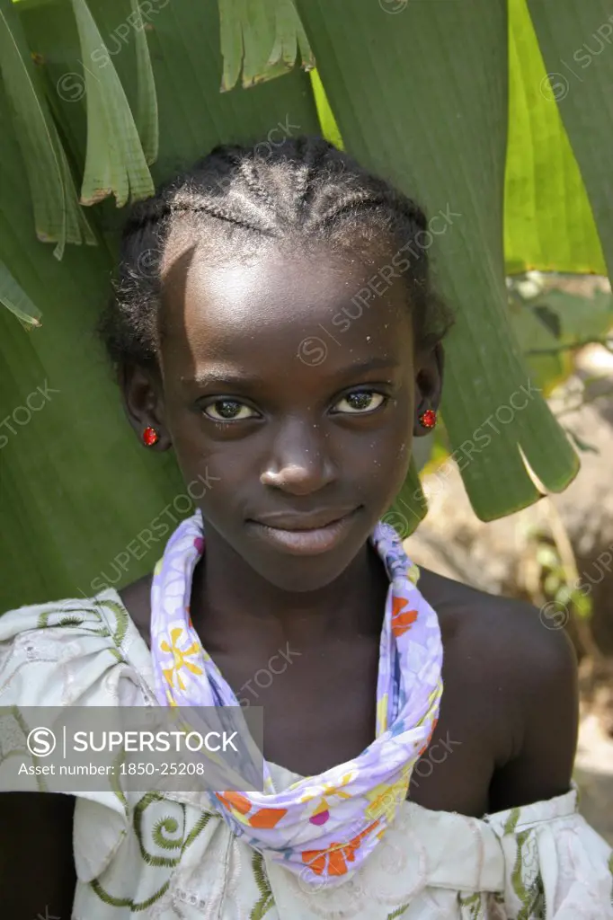 Gambia, Western Gambia, Tanji, Tanji Village.  Head And Shoulders Portrait Of Smiling Young Girl With Braided Hair And Almond Shaped Eyes Wearing Patterned Scarf Tied Around Her Neck And Red Earrings.