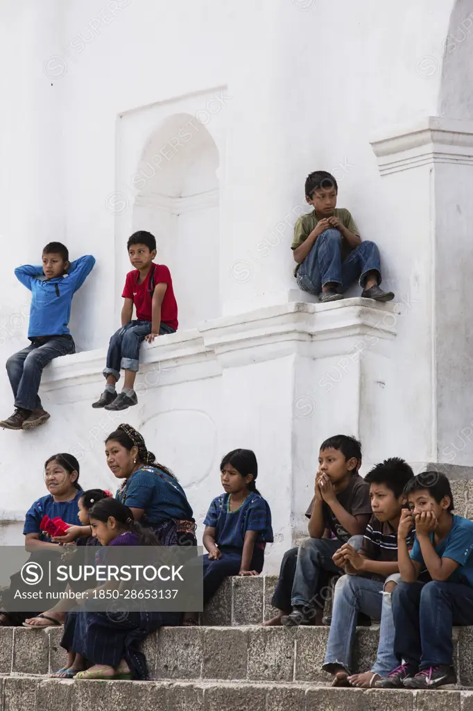 Guatemala, Solola Department, San Antonio Palopo, A Cakchiquel Mayan woman and children watch a program from the steps of the church. The woman and girls wear the traditional dress of San Antonio Palopo Guatemala, including the elaborate cinta or hair wrap, woven blue huipil blouse, faja or belt, and corte skirt.  The boys all wear modern clothing.