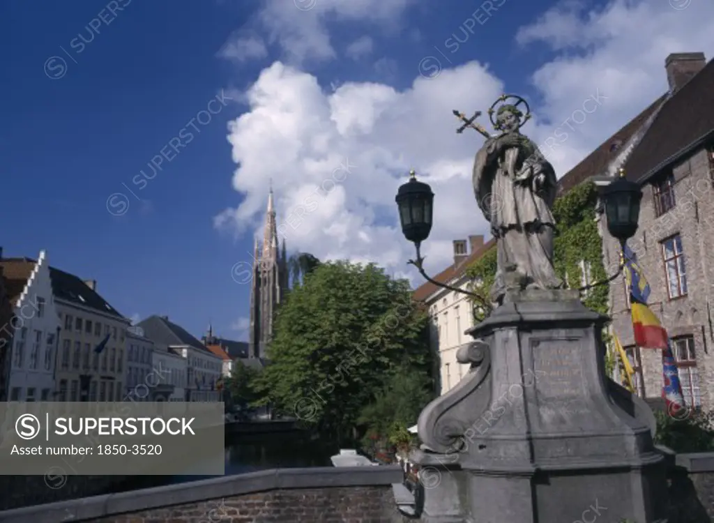 Belgium, West Flanders, Bruges, Religious Statue On Bridge Over Canal With Church Spire In Distance