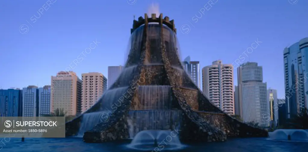 Uae, Abu Dhabi, The Volcano Fountain. Large Tiered Fountain With Water Cascading From Circular Top And High Rise Modern Buildings Behind.