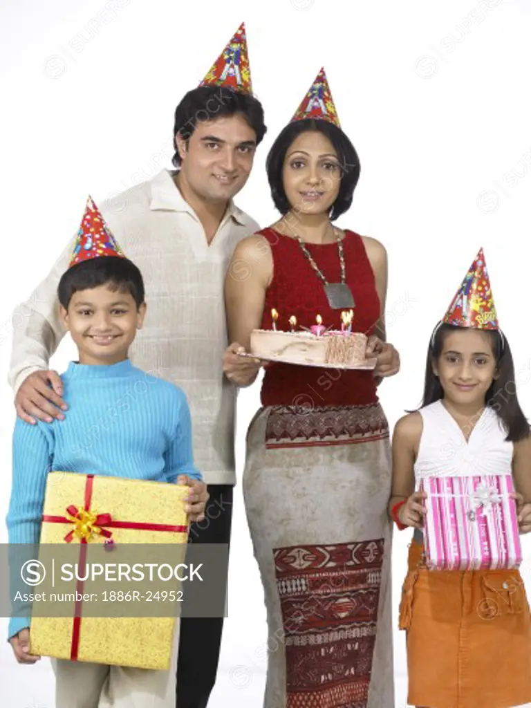 VDA200236 : South Asian Indian family with father, mother, son and daughter smiling carrying packet and birthday cake after shopping and looking at camera wearing dunce cap on head blue t-shirt, white pant, white and orange dress and cream color t-shirt and black pant, red dress, MR # 698, 699, 700, 701