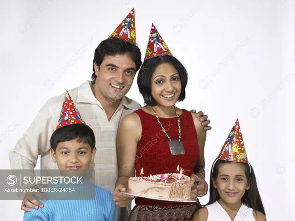 VDA200238 : South Asian Indian family with father, mother, son and daughter smiling carrying birthday cake after shopping and looking at camera wearing dunce cap on head blue t-shirt, white dress and cream color t-shirt, red dress, MR # 698, 699, 700, 701