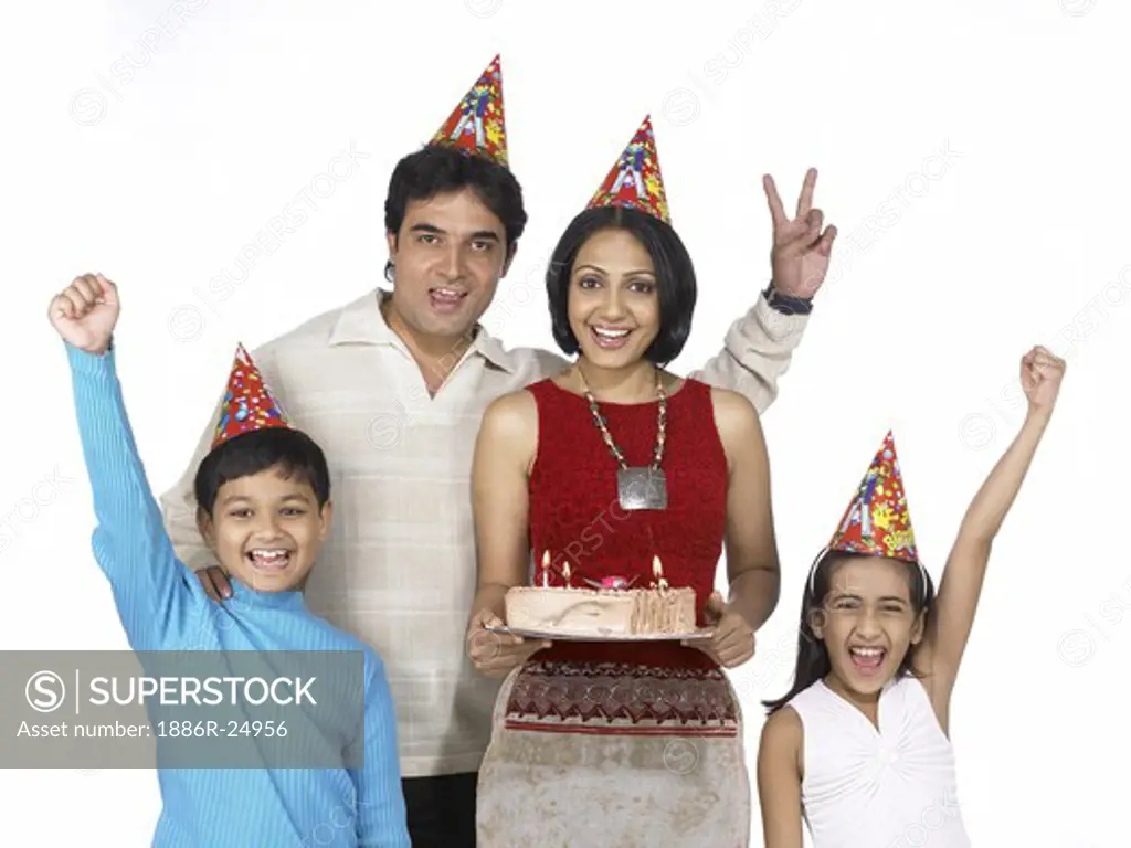 VDA200241 : South Asian Indian family with father, mother, son and daughter smiling carrying birthday cake after shopping and looking at camera wearing dunce cap on head blue t-shirt, white dress and cream color t-shirt, red dress, MR # 698, 699, 700, 701