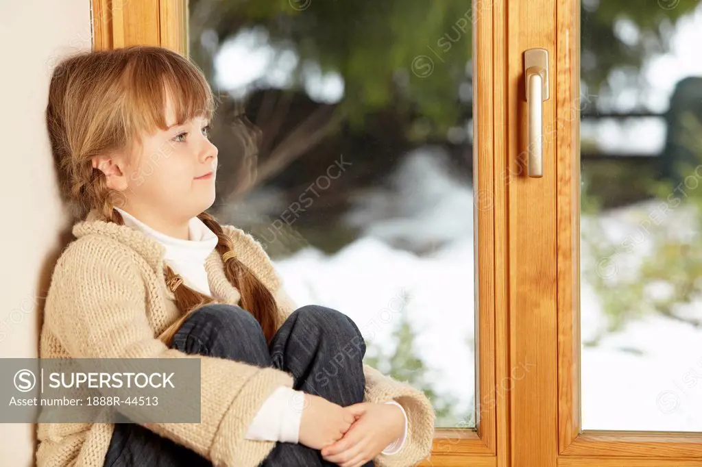 Young Girl Sitting On Window Ledge Looking At Snowy View