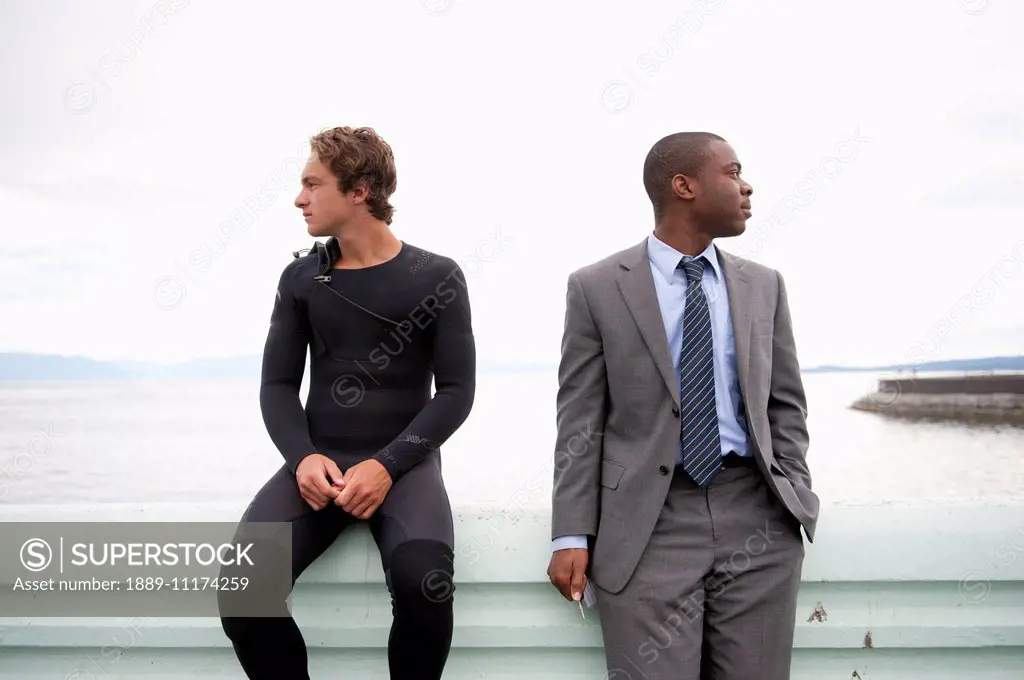 Two men of different ethnicities sitting on a ledge at the water's edge looking in different directions; Victoria, British Columbia, Canada