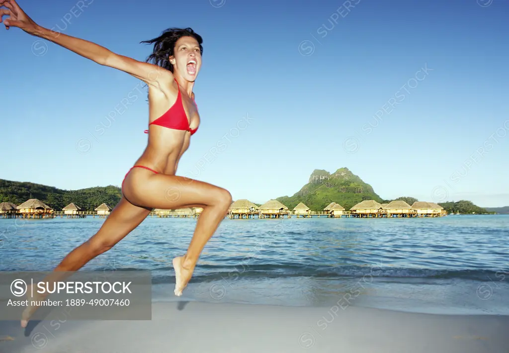 French Polynesia, Tahiti, Bora Bora, Woman Leaping On The Shore With Bungalows In The Background.