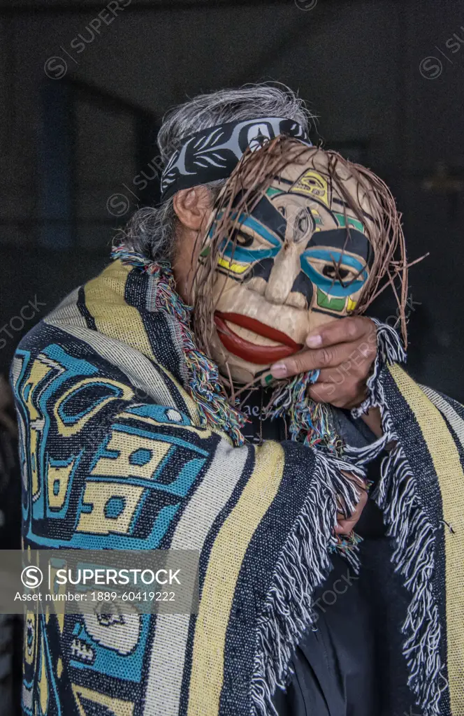 Man stands draped in a blanket with Native Indian design and holding a mask to obscure his face; Hoonah, Alaska, United States of America