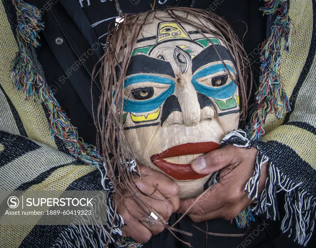 Man stands draped in a blanket with Native Indian design and holding a mask; Hoonah, Alaska, United States of America