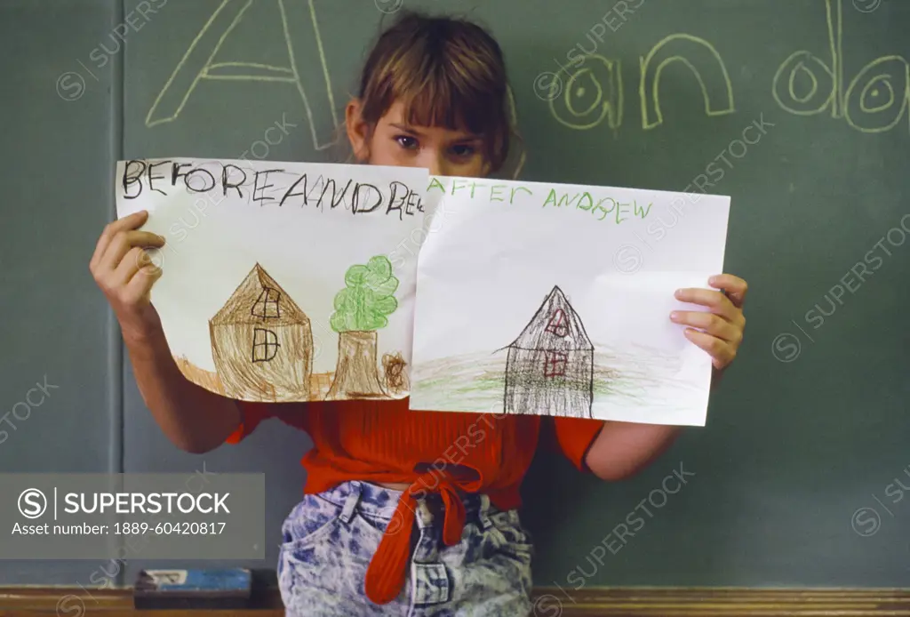 Young girl in a classroom holds up before and after drawings of her house after Hurricane Andrew destroyed it; Florida, United States of America