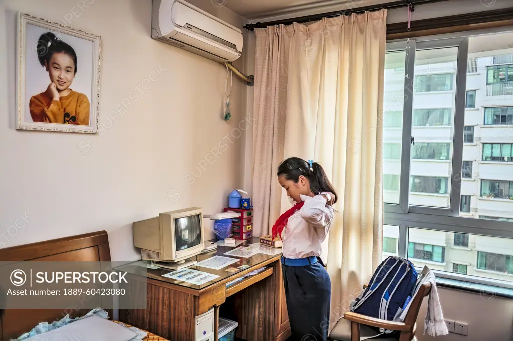 Teenage schoolgirl adjusts her uniform at a desk with a computer. Her portrait hangs on the wall above her; Shanghai, People's Republic of China