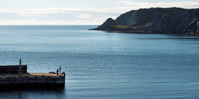 Rugged cliffs along the atlantic coastline and two people standing on a pier in the foreground; Twillingate, Newfoundland and Labrador, Canada