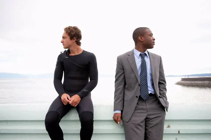 Two men of different ethnicities sitting on a ledge at the water's edge looking in different directions; Victoria, British Columbia, Canada