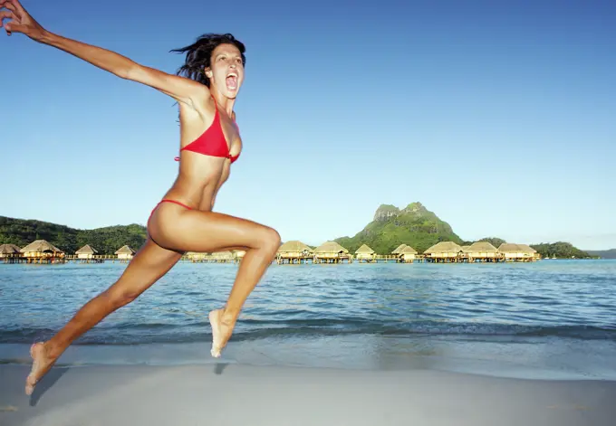 French Polynesia, Tahiti, Bora Bora, Woman Leaping On The Shore With Bungalows In The Background.