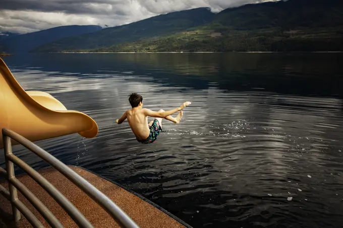 Boy jumping out of the end of a waterslide on a houseboat on a fall day on Shuswap Lake; British Columbia, Canada