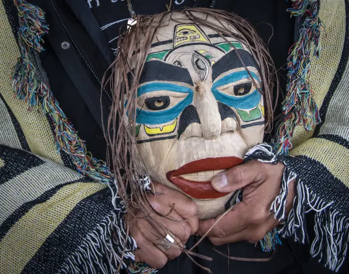 Man stands draped in a blanket with Native Indian design and holding a mask; Hoonah, Alaska, United States of America