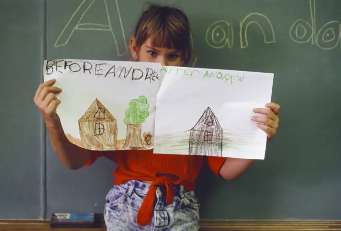 Young girl in a classroom holds up before and after drawings of her house after Hurricane Andrew destroyed it; Florida, United States of America