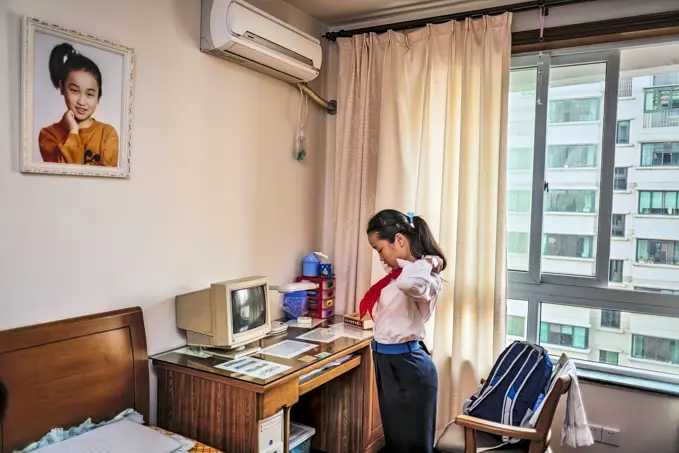 Teenage schoolgirl adjusts her uniform at a desk with a computer. Her portrait hangs on the wall above her; Shanghai, People's Republic of China