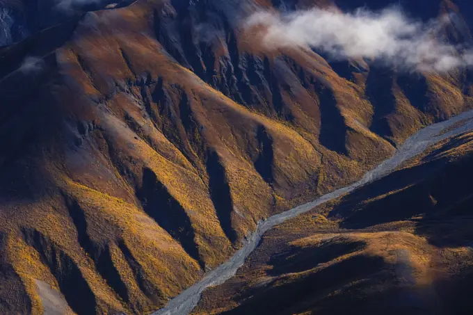 Late afternoon sun lights up the autumn colors on the slopes as Nines Creek flows through the mountains on its way to the Arctic Ocean; Destruction Bay, Yukon, Canada