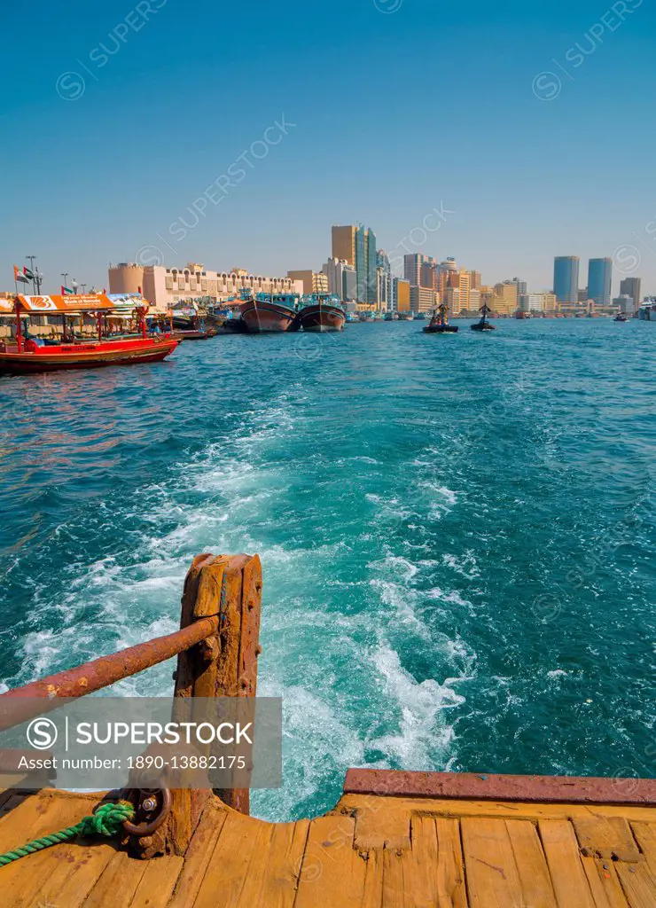 Modern Dubai cityscape disappears in the wake of an old water taxi speeding past the dhow harbour up Dubai Creek, Dubai, United Arab Emirates, Middle ...