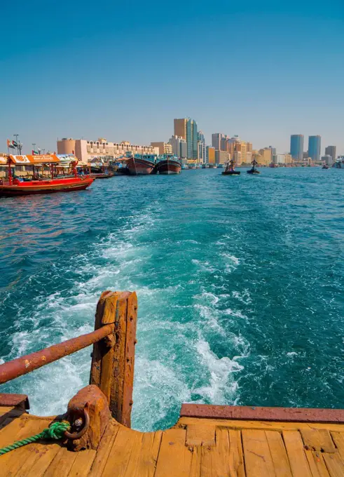 Modern Dubai cityscape disappears in the wake of an old water taxi speeding past the dhow harbour up Dubai Creek, Dubai, United Arab Emirates, Middle ...