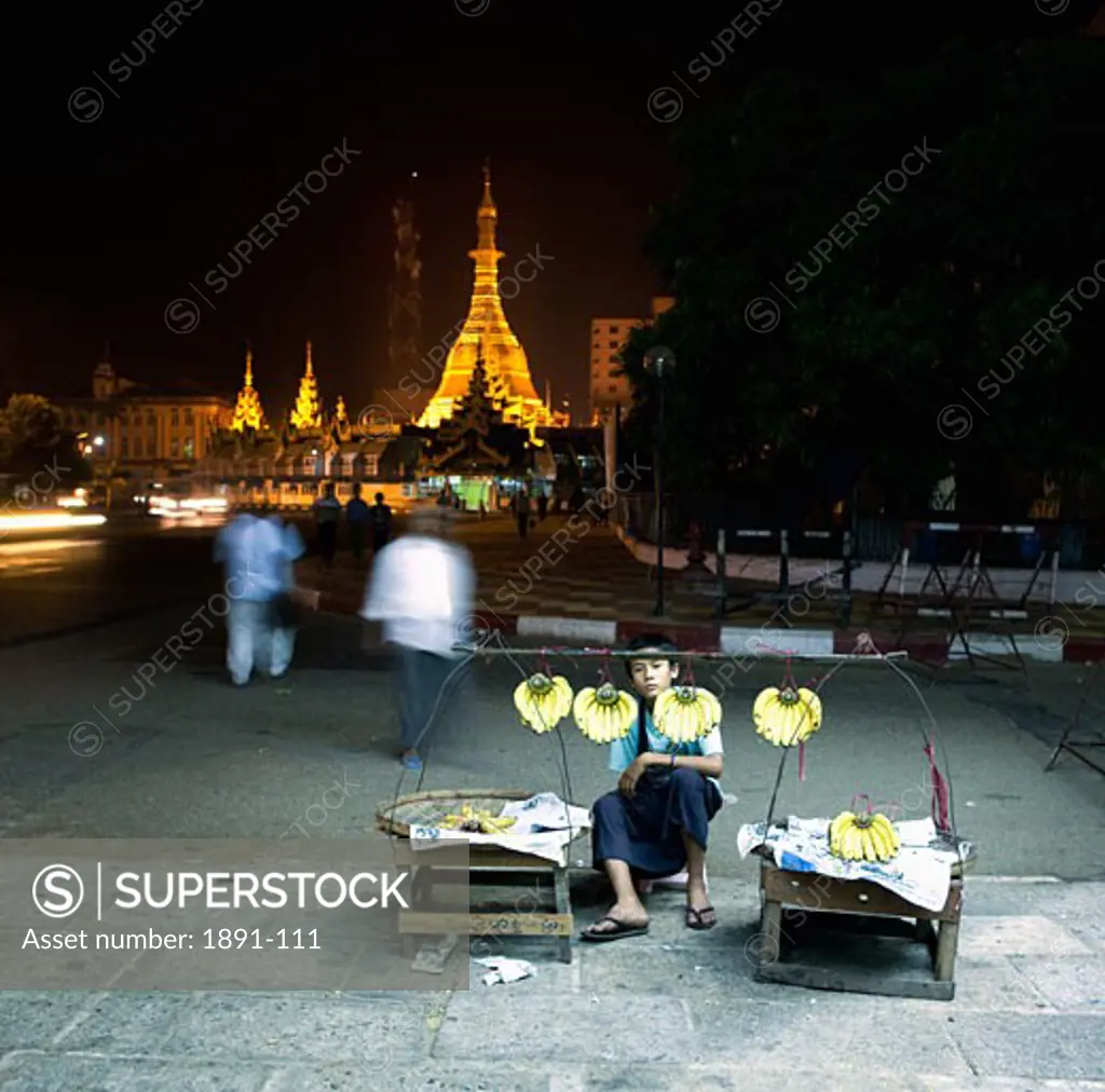 Teenage boy selling bananas with a pagoda lit up in the background, Yangon, Myanmar