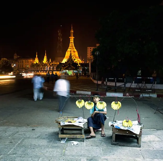 Teenage boy selling bananas with a pagoda lit up in the background, Yangon, Myanmar