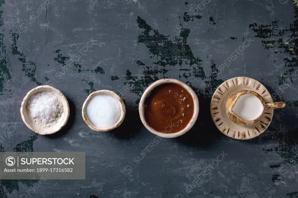 Homemade salted caramel sauce with grains of fleur de sel salt. Ingredients in ceramic bowls in row. Grey textured canvas background. Flat lay. space