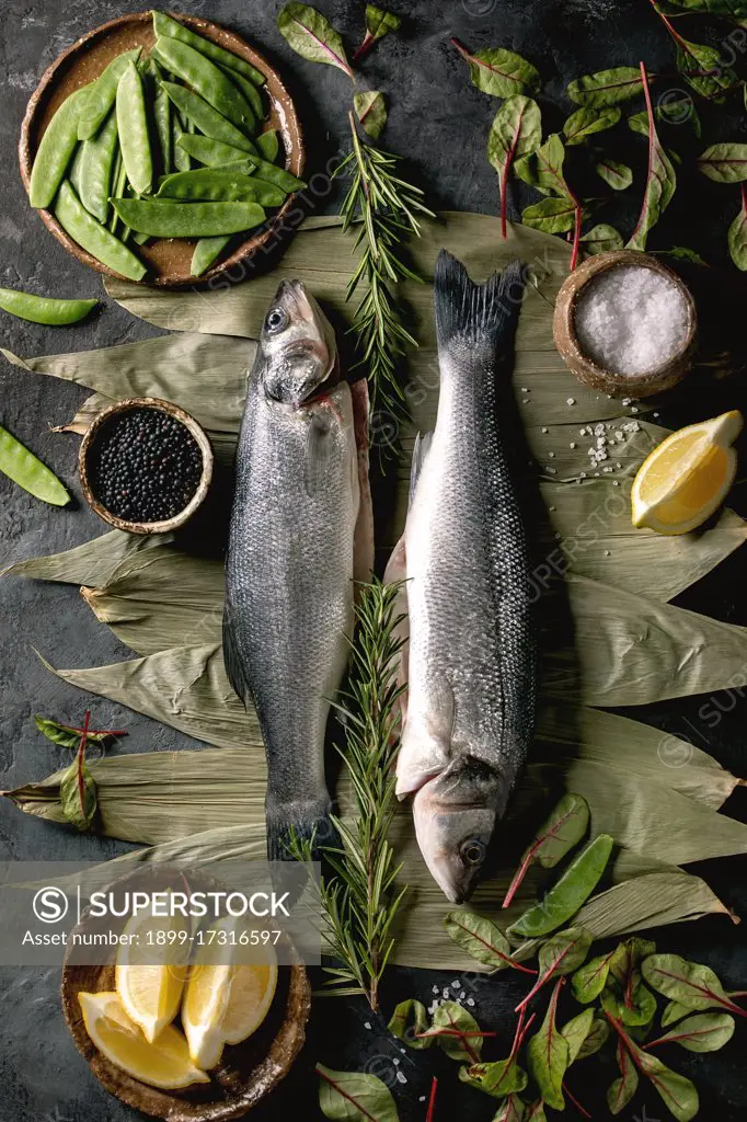 Raw fresh uncooked fish sea bass on dry bamboo leaves ready to cook with lemon. sea salt. black and green beans. green salad over dark texture concrete background. Flat lay. space