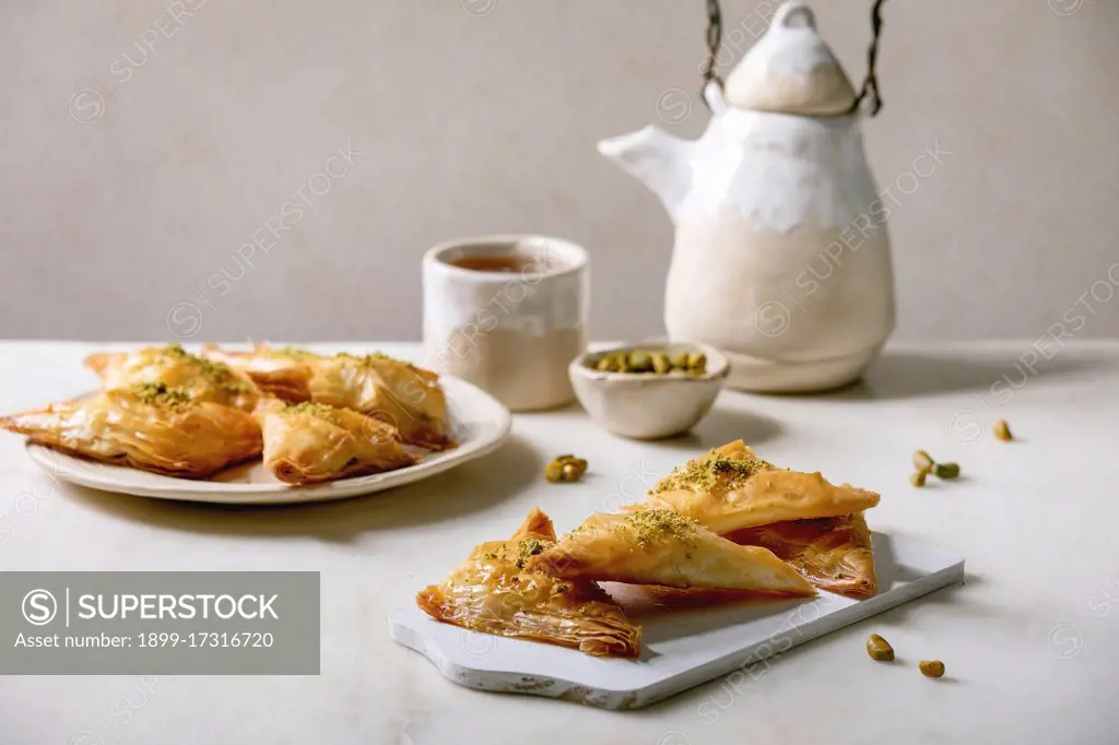 Homemade Turkish traditional dessert baklava with pistachio served on white wooden cutting board with bowl of nuts. cup of tea. ceramic teapot over white marble table.