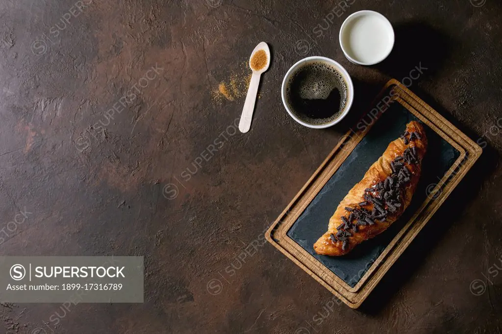 Traditional french puff pastry and chocolate. croissant on slate wooden board with paper cups of coffee and milk. recycled wooden spoon of cane sugar over dark texture background. Flat lay. space