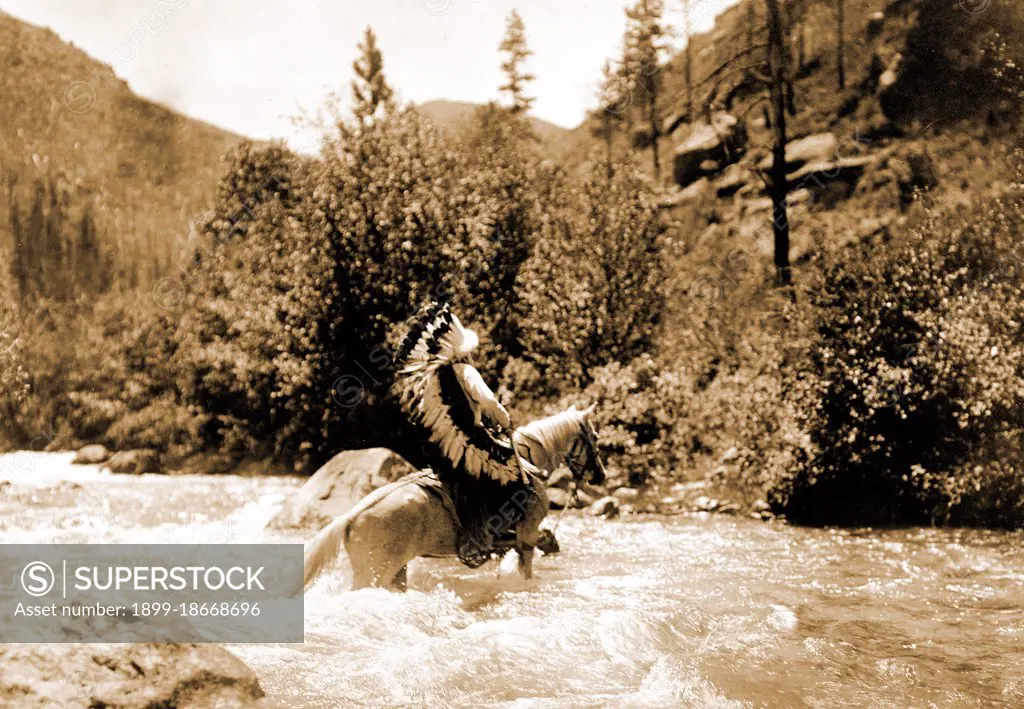 Edward S. Curtis Native American Indians - Bullchief, wearing warbonnet, crossing shallow rapids on horseback circa 1905.