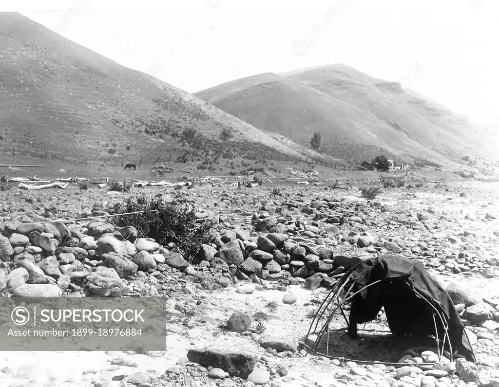 Landscape, lashed pole framed with blanket, large smooth stones in dry river bed, house, fence and hills in background. 