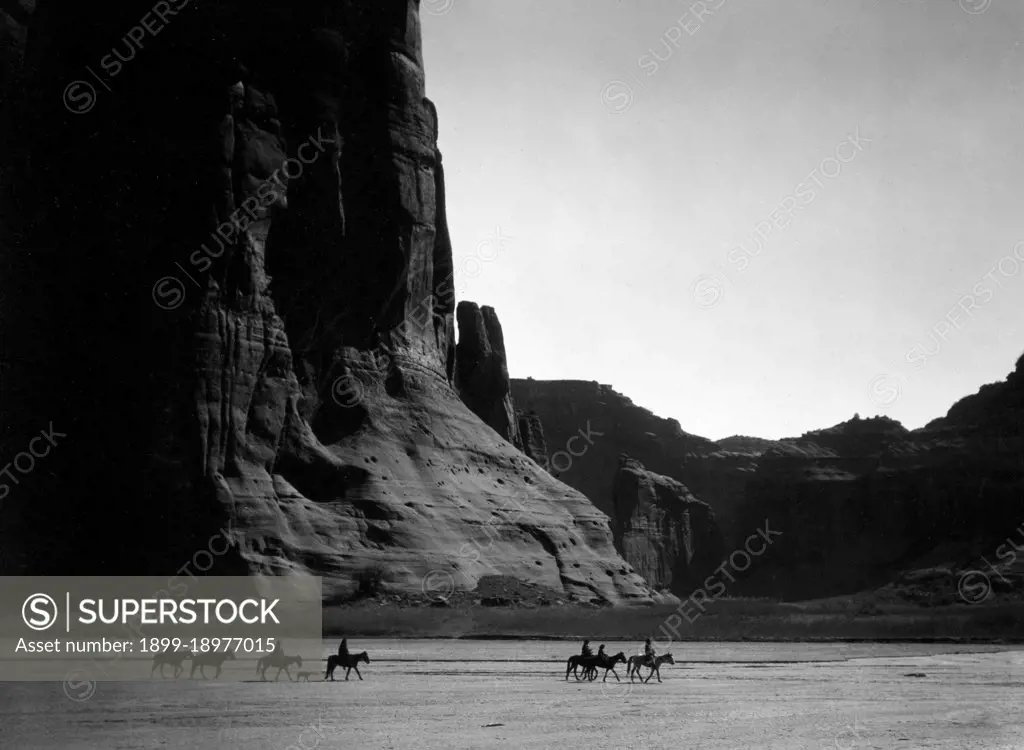 Seven Navajo riders on horseback and dog traveling against a background of canyon cliffs. 