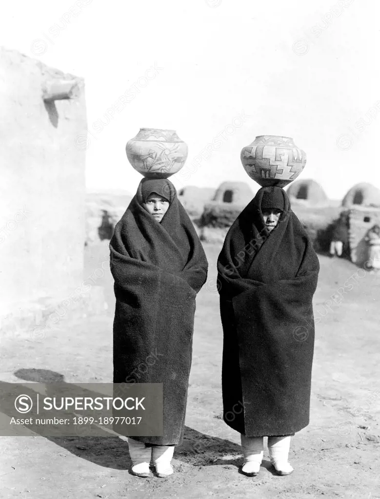 Two females, full length, wearing blankets, pots on heads. 