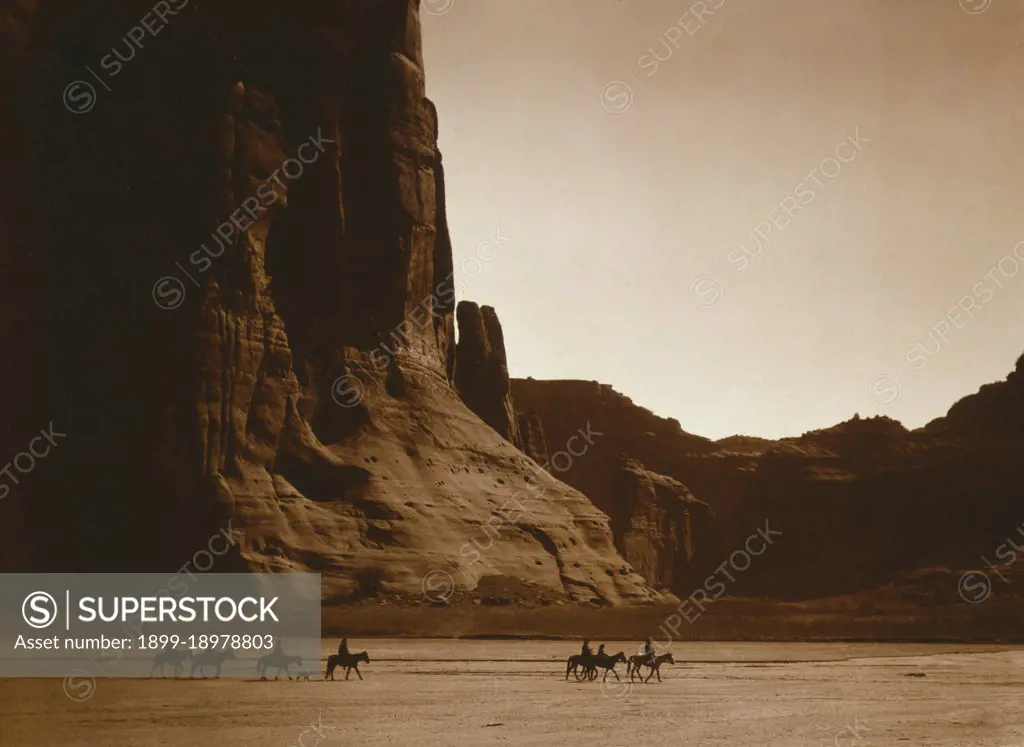 Seven Navajo riders on horseback and dog traveling against a background of canyon cliffs. 