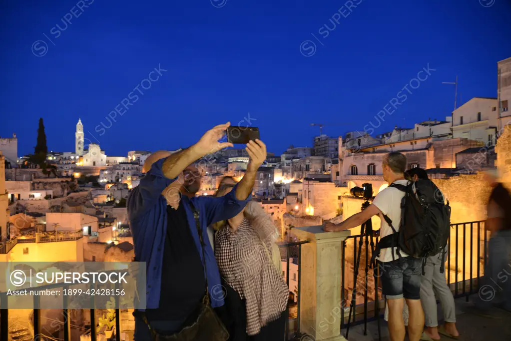 Tourist taking pictures and selfies. At night with suggestive lights the Pontifical Basilica Cathedral of Maria Santissima della Bruna and Sant'Eustachio dominates the view over Matera. Matera is a city located on a rocky outcrop. The so-called area of the Sassi (Stones) is a complex of cave houses carved into the rock, Basilicata, Italy, Europe