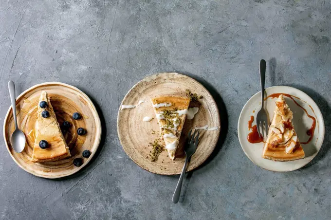 Pieces of cheesecake with different toppings and decor on ceramic plates with forks in row over grey texture background. Flat lay. space