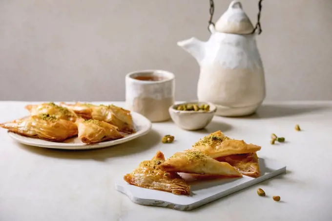 Homemade Turkish traditional dessert baklava with pistachio served on white wooden cutting board with bowl of nuts. cup of tea. ceramic teapot over white marble table.