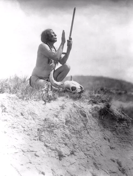 Edward S. Curits Native American Indians - Slow Bull, squatting, wearing breechcloth, holding pipe with mouthpiece pointing skyward, buffalo skull at his feet circa 1907.