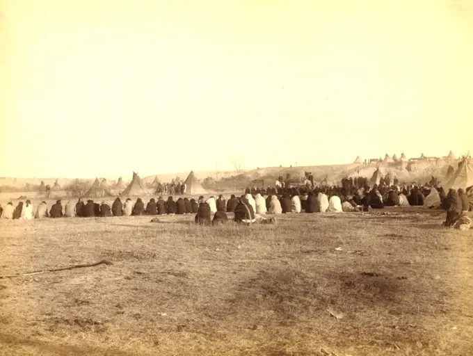 Rear view of a large semi-circle of Lakota men sitting on the ground, with tipis in background, probably on or near Pine Ridge Reservation. 1891.