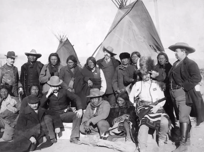  Group of White Americans and Lakota (Brulé, Miniconjou, and Oglala) men standing and sitting in two rows in front of tipis..