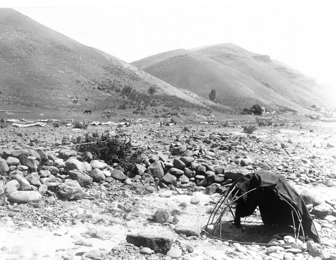 Landscape, lashed pole framed with blanket, large smooth stones in dry river bed, house, fence and hills in background. 