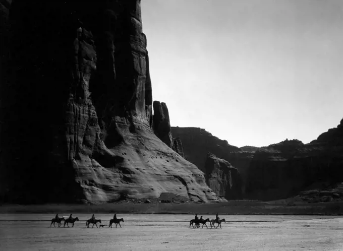 Seven Navajo riders on horseback and dog traveling against a background of canyon cliffs. 