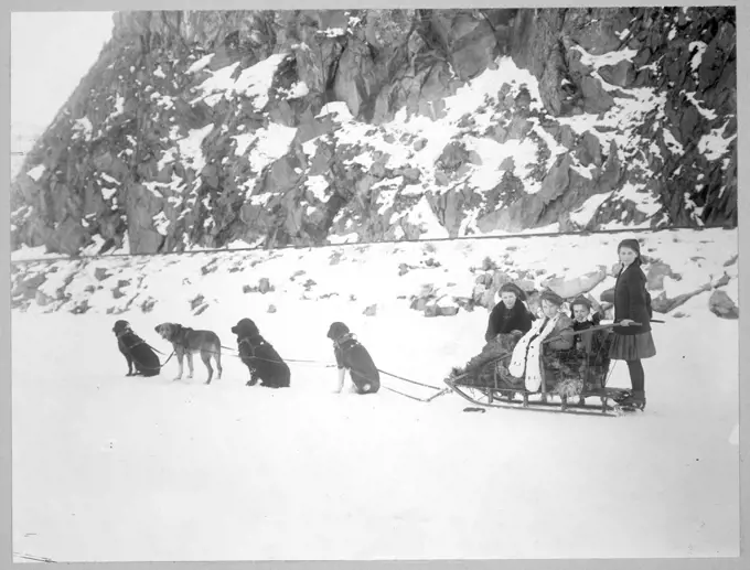 Dog team harnessed to a sled holding four girls, in front of a snow-covered cliff. 1900-1917 Canada. 