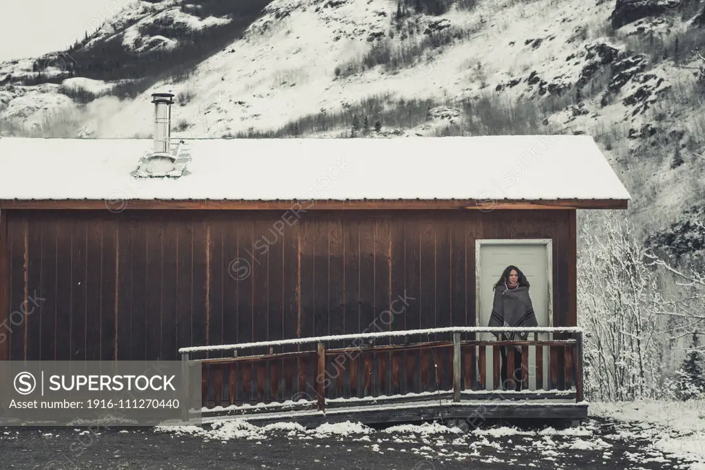 A young Woman is wrapped in a woolen blanket outside of a simple wooden cabin. Yukon Territory, Canada