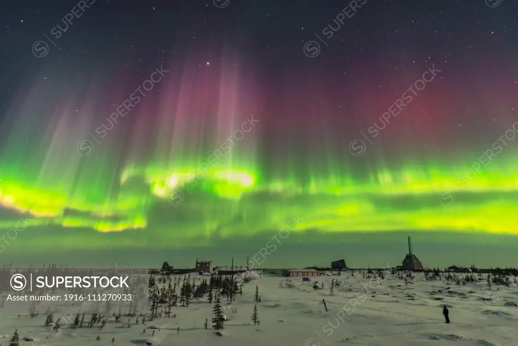 A superb display of aurora borealis seen on March 14, 2016 when it reached Level 5 storm levels. This was as the major substorm hit and the curtains to the south lit up with intensity and motion. Note the subtle differences in red coloration in the tops of the curtains. This was seen and shot from the Churchill Northern Studies Center, Churchill, Manitoba, with this image looking south over the abandoned Churchill Rocket Range. A lone observer admires the show. Illumination is from the 6-day o