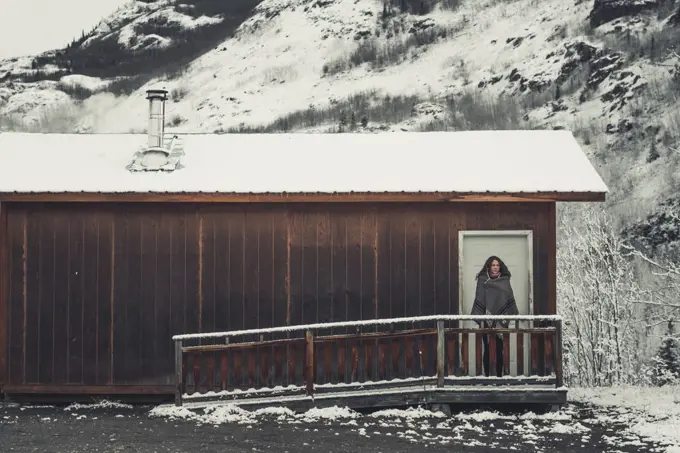 A young Woman is wrapped in a woolen blanket outside of a simple wooden cabin. Yukon Territory, Canada