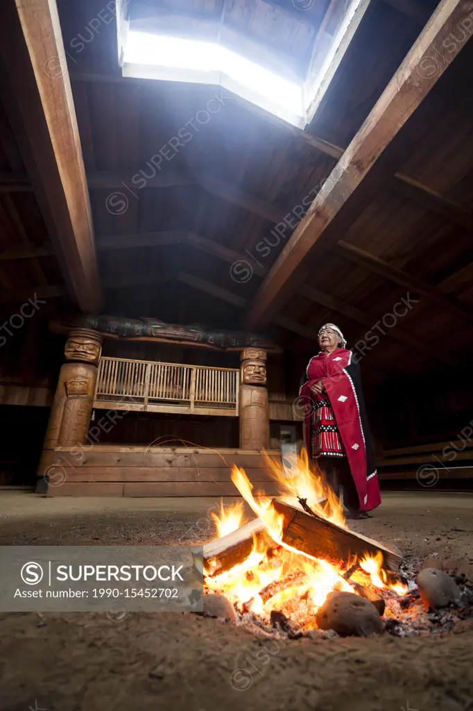 A first nations elder dressed in her traditional regalia stands beside a fire in the bighouse in the village of Oweekeno. Wuikinuxv, Rivers Inlet, Bri...