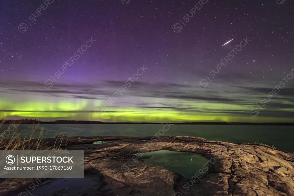 Aurora dancing and a meteor streaking over lake in Manitoba Canada big boulder in the eastman region with lcouds adding texture to a beautiful sky